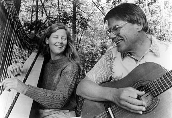 Photo of Carol playing harp and Gordon playing guitar in a forest.
