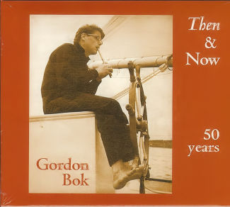 Image of Gordon Bok playing a pipe sitting on a sailboat. The boom and sail are visible in the background, and Gordon's bare foot is holding the boat's wheel.