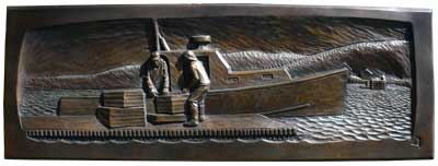 Photo of a bronze casting of a bas-relief carving of two fishermen unloading crates of their catch from a lobster boat tied to a dock, with hills in the background.